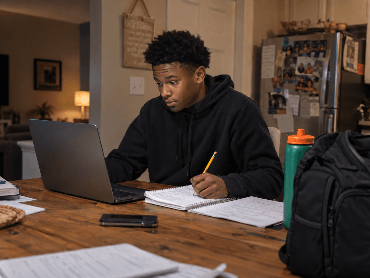 A student working with a notebook and laptop at home, with a sports bag and water bottle beside him.